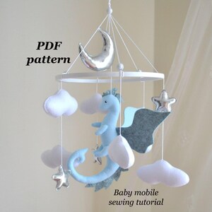 May include: A baby mobile featuring a light blue dragon, silver moon and stars, and white clouds. The mobile is suspended from a white circular frame. Text on the image reads "PDF pattern" and "Baby mobile sewing tutorial".