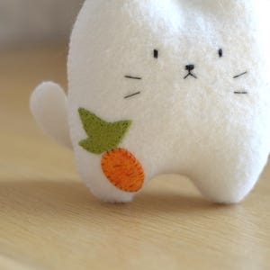 May include: A white felt bunny plush toy with a carrot applique. The bunny has a simple face with black eyes, a nose, and whiskers. The carrot is orange with a green leaf. The toy is on a wooden surface.