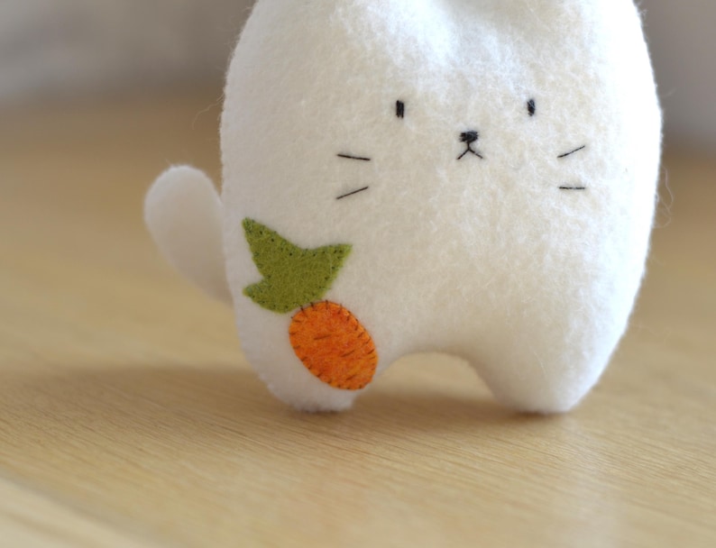 May include: A white felt bunny plush toy with a carrot applique. The bunny has a simple face with black eyes, a nose, and whiskers. The carrot is orange with a green leaf. The toy is on a wooden surface.