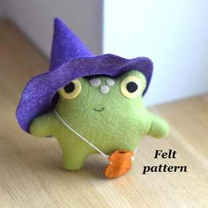 May include: A handmade felt frog doll in light green, wearing a purple witch hat and carrying an orange pouch. The frog has large yellow eyes and a smiling face. The text "Felt pattern" is visible.