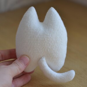 May include: A small, white, plush cat toy with a rounded body, two pointed ears, and a curved tail. The toy is made of a soft, textured material and is held in a person's hand. The background is a light brown wooden surface.