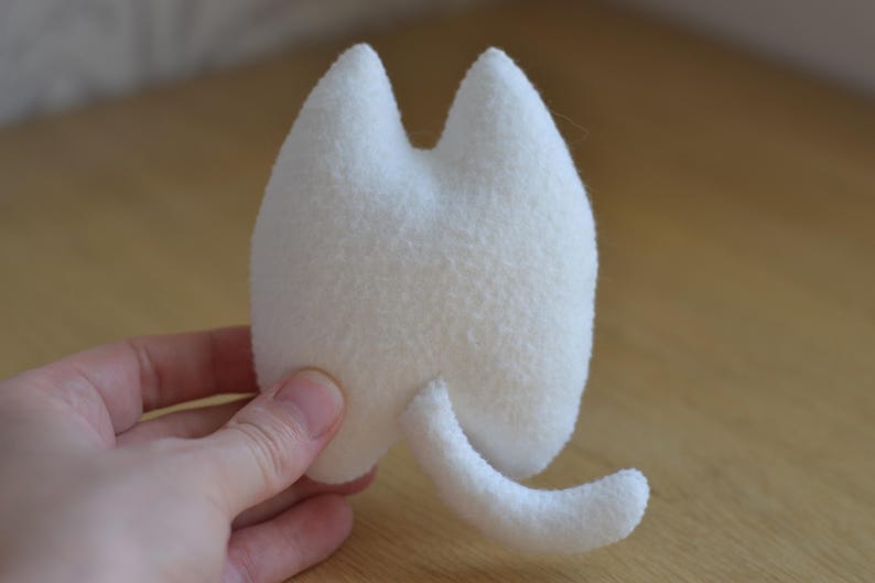 May include: A small, white, plush cat toy with a rounded body, two pointed ears, and a curved tail. The toy is made of a soft, textured material and is held in a person's hand. The background is a light brown wooden surface.