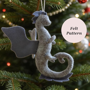 May include: A grey felt dragon ornament featuring a curved tail, small wings, and a crest along its back. The ornament hangs from a white string and includes a circular white tag with the black text Felt Pattern printed on the surface.