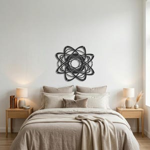 May include: A black metal wall sculpture with an abstract, geometric design, reminiscent of an atom or a stylized flower. The artwork is mounted on a white wall above a bed with neutral-toned bedding, creating a modern and minimalist aesthetic.