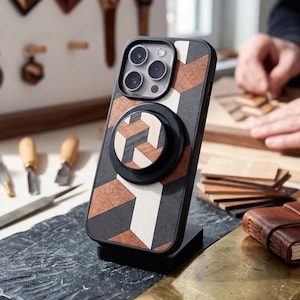 Op de afbeelding: Een houten telefoonhoesje met een geometrisch ontwerp in zwart, bruin en wit, met een bijpassende popsocket, staat op een zwarte standaard. Andere houten accessoires en gereedschappen op de achtergrond suggereren een ambachtelijk thema.