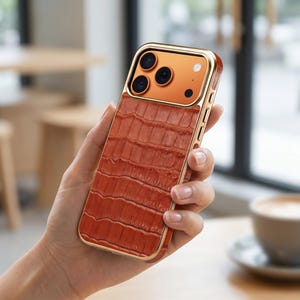 Puede incluir: Una funda de teléfono marrón con un marco dorado y un patrón texturizado similar a la piel de cocodrilo. La funda del teléfono tiene un módulo de cámara con tres lentes y un flash. La funda se sostiene en una mano.