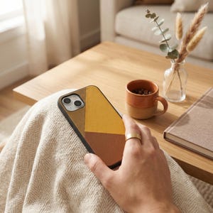 May include: A geometric phone case in shades of brown and yellow is held in a person's hand. The case has a black border and a textured appearance. A gold ring is on the person's finger. A small coffee cup and a book are on a wooden table.
