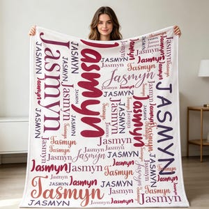 May include: A white blanket with the name "Jasmyn" printed in different sizes and shades of red. The name is repeated across the blanket in a decorative pattern. The blanket is held up by a person.