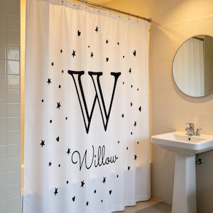 May include: A white shower curtain featuring a large black letter W and the name Willow in a cursive script, surrounded by a circular pattern of black stars, hearts, and dots. The curtain hangs from a silver rod with metal rings.