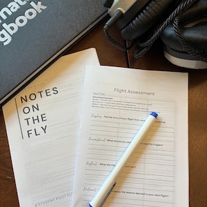 May include: Aviation-themed items on a wooden surface. Includes a black notebook with silver text, a headset, and printed pages titled "Notes on the Fly" and "Flight Assessment." A white pen with blue accents rests on the assessment sheet.