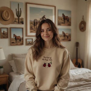 May include: A beige sweatshirt with the text "The last of the bugs club" and two red ladybug graphics. The sweatshirt is long-sleeved and has a crew neck. The image also shows framed art and a guitar.