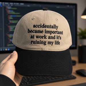 May include: A two-tone baseball cap featuring a beige crown and a black brim. The front of the crown displays black embroidered text that reads: accidentally became important at work and it's ruining my life. The cap has a structured six-panel design.