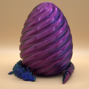 Könnte beinhalten: Ein großes, ovales Ei mit einer spiralförmigen, gerippten Textur in metallisch violetter Ausführung. Eine kleine, bewegliche Kreatur mit irisierenden blauen und violetten Schuppen ist um die Basis des Eies gewickelt und zeigt sichtbare segmentierte Gliedmaßen.