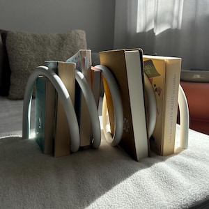 Könnte beinhalten: Ein weißer, gebogener Metall-Buchstütze hält eine Sammlung von Büchern mit verschiedenfarbigen Buchrücken. Die Buchstütze hat ein modernes Design mit einer Schlaufenform, die die Bücher umschließt. Die Bücher sind auf einer weichen, weißen Oberfläche angeordnet.