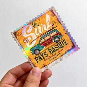 May include: A sticker with a vintage design featuring a teal and maroon station wagon with a surfboard on top. The sticker has the words "Le Surf" and "Pays Basque" with the text "Côte Atlantique - France Depuis 1960". The sticker has a holographic border.