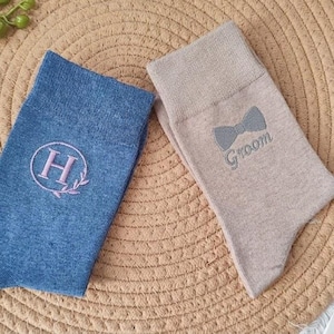 May include: Two pairs of socks, one blue and one beige, displayed on a woven mat. The blue sock has a pink embroidered letter "H" inside a circle with a floral design. The beige sock features a bow tie and the word "Groom".