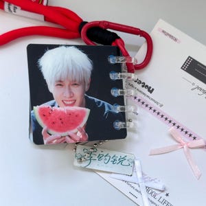 Peut inclure: Un mini carnet carré présentant la photo d'une personne aux cheveux blancs tenant une tranche de pastèque. Le carnet est attaché à un cordon rouge avec un mousqueton et comprend une petite breloque rectangulaire avec des caractères chinois.