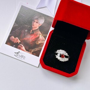 May include: A silver ring featuring two textured wing-shaped sides, a central square-cut red gemstone, and a small round clear stone, presented in a red velvet box next to a rectangular card showing an illustration of a person with silver hair.