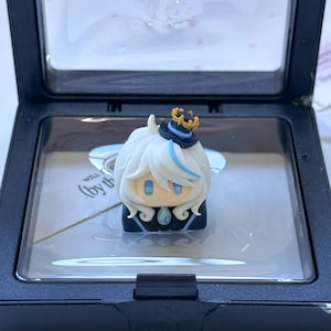 May include: A small, sculpted character figurine with white hair, a blue streak, and a black top hat with a gold crown detail. The figure has blue eyes and a light blue teardrop-shaped gem on its chest, displayed inside a black plastic case.