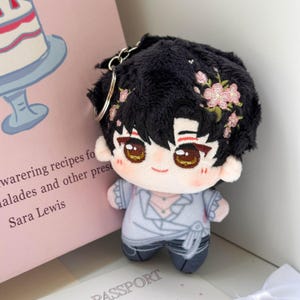 May include: A plush doll keychain featuring a character with black hair adorned with pink floral embroidery, large brown eyes, and a light complexion. The doll wears a light blue wrap-style top with ruffled sleeves and dark blue pants, attached to a silver metal ring.