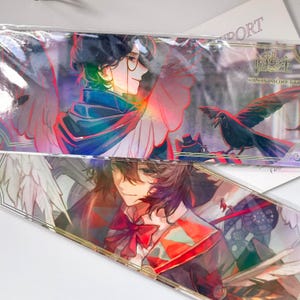 May include: Two rectangular bookmarks featuring anime-style illustrations. The top bookmark shows a character with dark hair, glasses, and white wings, alongside a black crow. The bottom bookmark depicts a character with brown hair and a red bow tie.