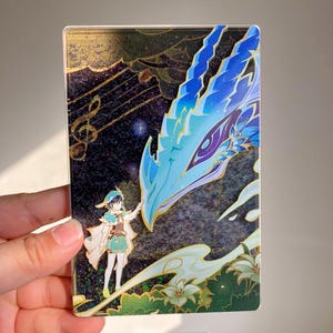 Op de afbeelding: Een rechthoekig acrylkunstwerk met een personage in groen en wit dat interactie heeft met een blauwe draak. De achtergrond heeft muzieknoten en een sterrennachtontwerp met gouden accenten. Het kunstwerk wordt door een hand vastgehouden.