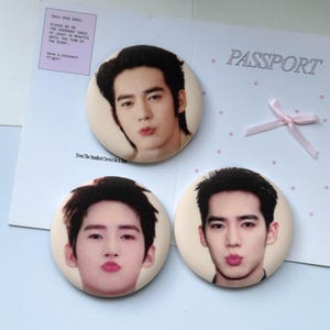 Könnte beinhalten: Drei runde Ansteckbuttons mit dem Porträt einer Person mit dunklem Haar und gespitzten Lippen. Die Buttons liegen auf einem weißen Papierhintergrund, der mit dem Wort PASSPORT, einer kleinen rosa Schleife und Text wie PACK YOUR BAGS bedruckt ist.