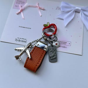 May include: A keychain featuring a silver airplane charm, a textured brown leather loop, a red apple-shaped ring, a small white heart charm with a red center, and a rectangular metal tag engraved with the text "When U come back".