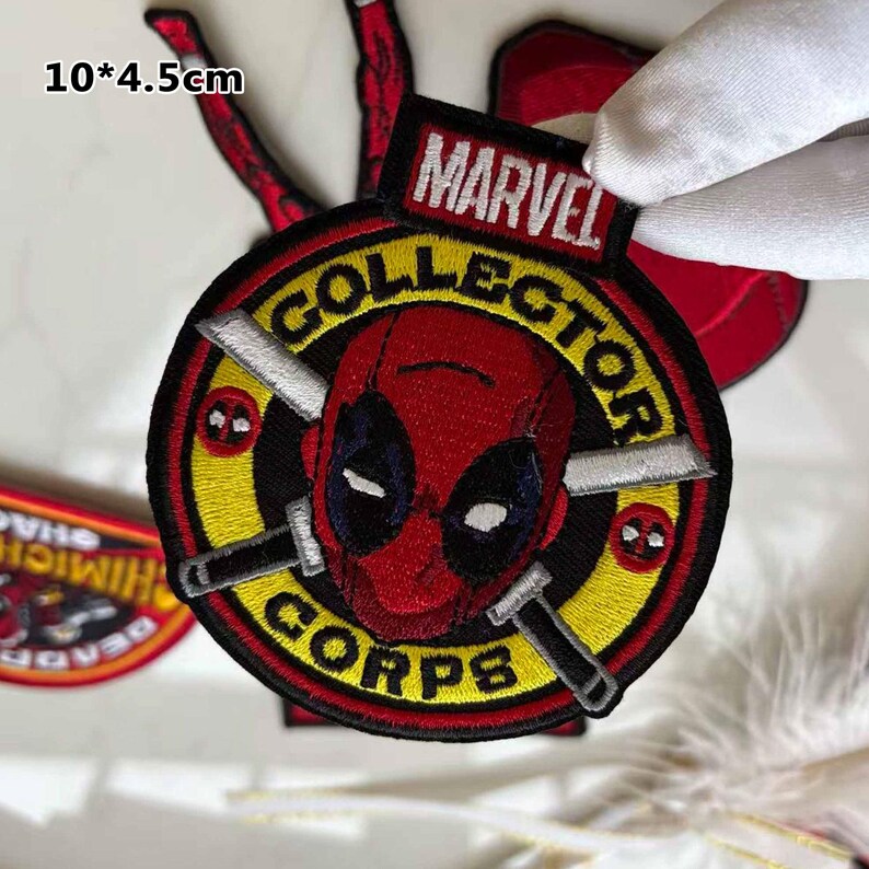 May include: Embroidered patch featuring the Deadpool character from Marvel Comics. The patch is round, with a red and black design, and the text "MARVEL COLLECTOR CORPS". The patch measures 10 x 4.5 cm.