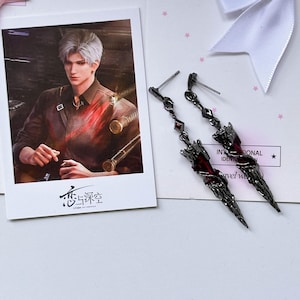 May include: A pair of long, dangling earrings featuring a dark metallic finish and deep red, jagged, crystalline-like accents. The earrings are displayed next to a rectangular card showing an illustration of a person with silver hair.