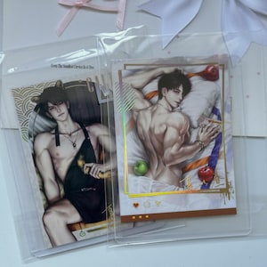 May include: Two rectangular art prints in clear plastic sleeves. The left print shows a person with cat ears wearing a black apron. The right print depicts a person lying on a white pillow with a green apple and a red apple, featuring gold foil accents.