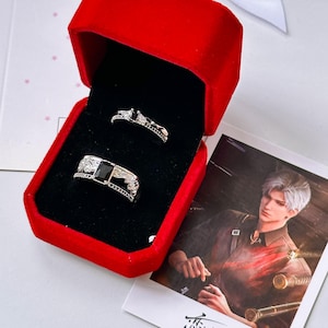 May include: A pair of silver-toned rings with black rectangular stones, featuring intricate side engravings, presented inside a bright red velvet jewelry box with a black interior lining, accompanied by a printed photograph of a person.