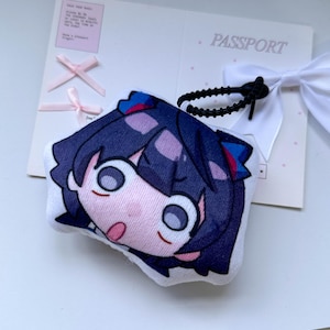 May include: A plush keychain featuring a stylized anime character head with dark blue hair, large eyes, and an open mouth expression. The plush is attached to a black coiled plastic wrist strap and rests on a white card printed with the word PASSPORT.