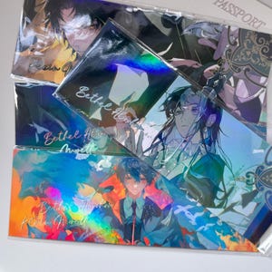 May include: A collection of rectangular holographic prints featuring anime-style character illustrations. Each print displays vibrant colors, intricate line art, and the text Bethel Abraham Klein Moretti in cursive script, all encased in clear plastic sleeves.