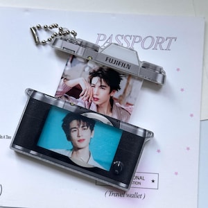 May include: A silver and black Fujifilm camera-shaped keychain with a metal ball chain. The camera body features a rectangular display window showing a portrait of a person, with a second portrait visible in the top section. The design includes the text FUJIFILM.