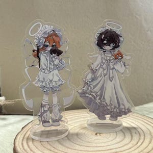 Puede incluir: Dos figuras acrílicas de personajes de anime. Una figura tiene el pelo naranja y sostiene un pequeño peluche negro. La otra figura tiene el pelo oscuro y sostiene un pequeño peluche naranja. Ambas figuras visten trajes blancos con temática de ángeles.