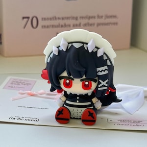 May include: A small, stylized figurine of a character with dark hair, red eyes, and a white headpiece, wearing a black and white outfit with red accents on the feet. The figure is positioned on a white card with text including by the moon.