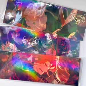 May include: Three rectangular bookmarks featuring holographic anime-style illustrations. The top shows a character with light hair in green, the middle a dark-haired figure in red, and the bottom a red-haired character. Each includes text and star accents.