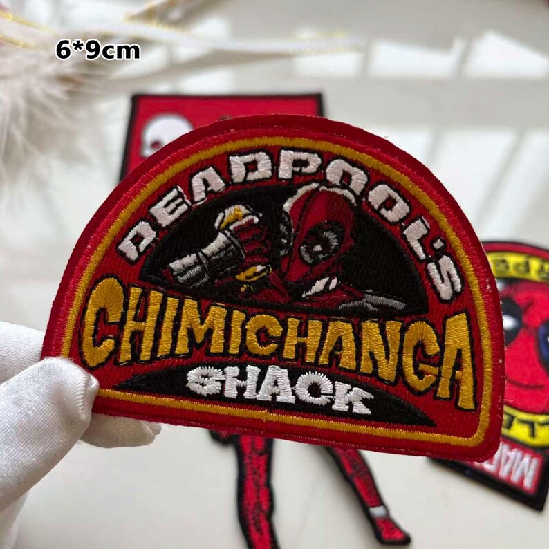 May include: A red and black embroidered patch with the text "DEADPOOL'S CHIMICHANGA SHACK" in yellow. The patch features a Deadpool character design. The patch measures 6 x 9 cm.