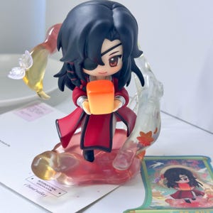 Puede incluir: Una figura de anime estilo chibi con cabello negro y un parche en el ojo, vistiendo un atuendo rojo. La figura sostiene un objeto rectangular naranja brillante, rodeado de elementos translúcidos con una pequeña mariposa blanca y hojas.