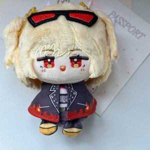 May include: A plush doll with blonde hair, red eyes, and black sunglasses resting on its head. The doll wears a dark jacket with a red flame pattern along the hem, a striped shirt, and yellow-accented shoes. The doll has embroidered facial features.
