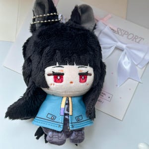 May include: A plush doll featuring black hair, pointed ears with a silver ball chain, and red embroidered eyes. The doll wears a bright blue jacket with a yellow and purple accent strip, a white shirt, and dark gray pants. The doll has a pale face.