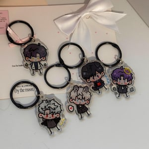 May include: Five acrylic keychains featuring cartoon characters with distinct hair colors and outfits, each attached to a black metal ring. One keychain includes a small pink ribbon. The characters have expressive faces and are rendered in a flat, colorful style.