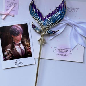 May include: A gold-toned metal hair stick featuring a decorative mermaid tail fin with blue and purple enamel inlay, positioned next to a rectangular card with a printed illustration of a person in a suit and the text "International Identification".