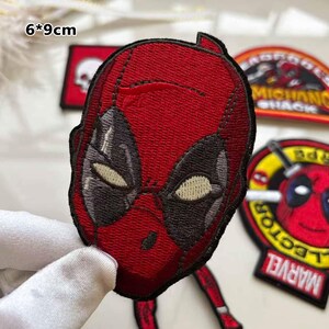May include: Embroidered Deadpool face patch, measuring 6 x 9 cm. The patch features a red mask with black eye and mouth details. Other patches with text and graphics are visible in the background.
