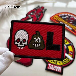 May include: A red rectangular patch with a black border featuring a white skull, a smiling poop emoji, and the letter "L". Other patches with comic book characters are visible.