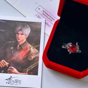 May include: A silver-toned ring featuring a central red heart-shaped stone surrounded by metallic wing-like accents and small pink crystals, presented inside a red velvet-lined box next to a character card with the text 恋与深空.