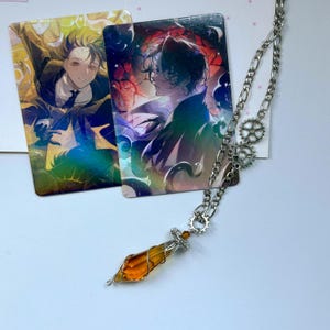 May include: A silver-toned chain necklace featuring two small gear-shaped charms and a faceted amber-colored crystal pendant wrapped in thin wire. Two rectangular trading cards with anime-style illustrations of characters are positioned behind the necklace.