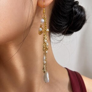 Peut inclure: Une longue boucle d'oreille pendante dorée composée de plusieurs chaînes délicates, de petites perles blanches et de breloques dorées en forme de coquillage et d'étoile de mer. Elle est suspendue à un crochet doré et inclut une perle en goutte.