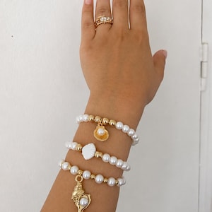 Peut inclure: Trois bracelets de perles composés de fausses perles blanches et de perles sphériques dorées. Le bracelet supérieur porte une breloque coquillage dorée avec une perle, le milieu un coquillage blanc, et le bas une grande conque dorée avec une perle.